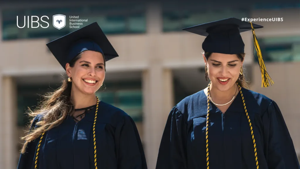 Two UIBS graduates wearing navy academic gowns and mortarboards during a graduation ceremony. The students are smiling outdoors, with gold honour cords and elegant accessories visible, while the UIBS logo and #ExperienceUIBS branding appear at the top of the image.
